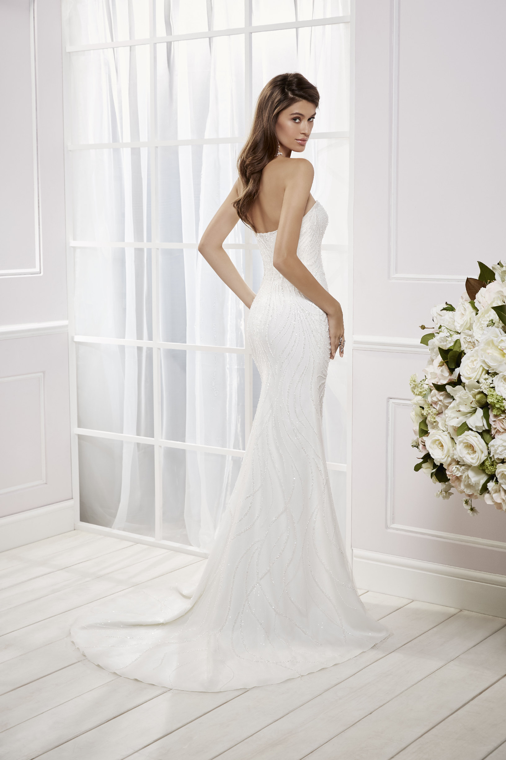 Brunette model stood in a white room by cream flowers in Ronald Joyce 69465, a heavily beaded strapless sweetheart fit and flare wedding dress with ivory beads and a puddle train