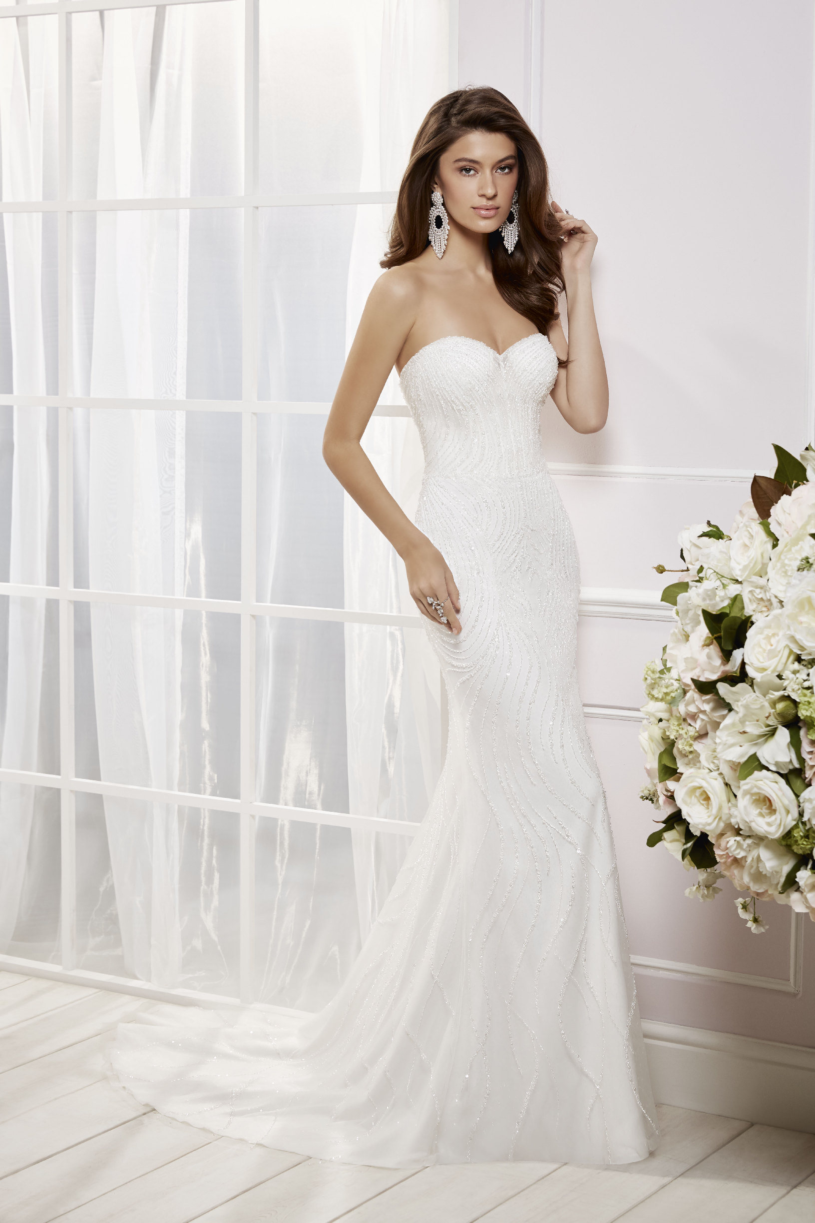 Brunette model stood in a white room by cream flowers in Ronald Joyce 69465, an exquisitely beaded strapless sweetheart fit and flare wedding dress with ivory beads