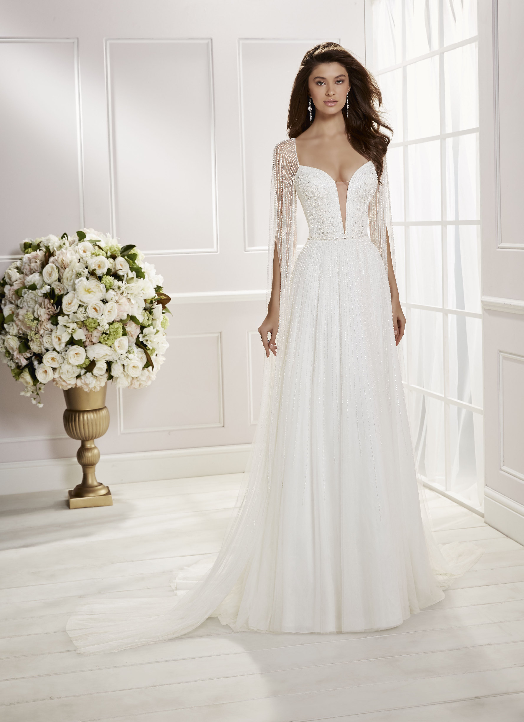 Brunette model stood in a white room by cream flowers in Ronald Joyce 69467, an ivory beaded A-line wedding dress with illusion beaded cape sleeves and a deep plunging neckline