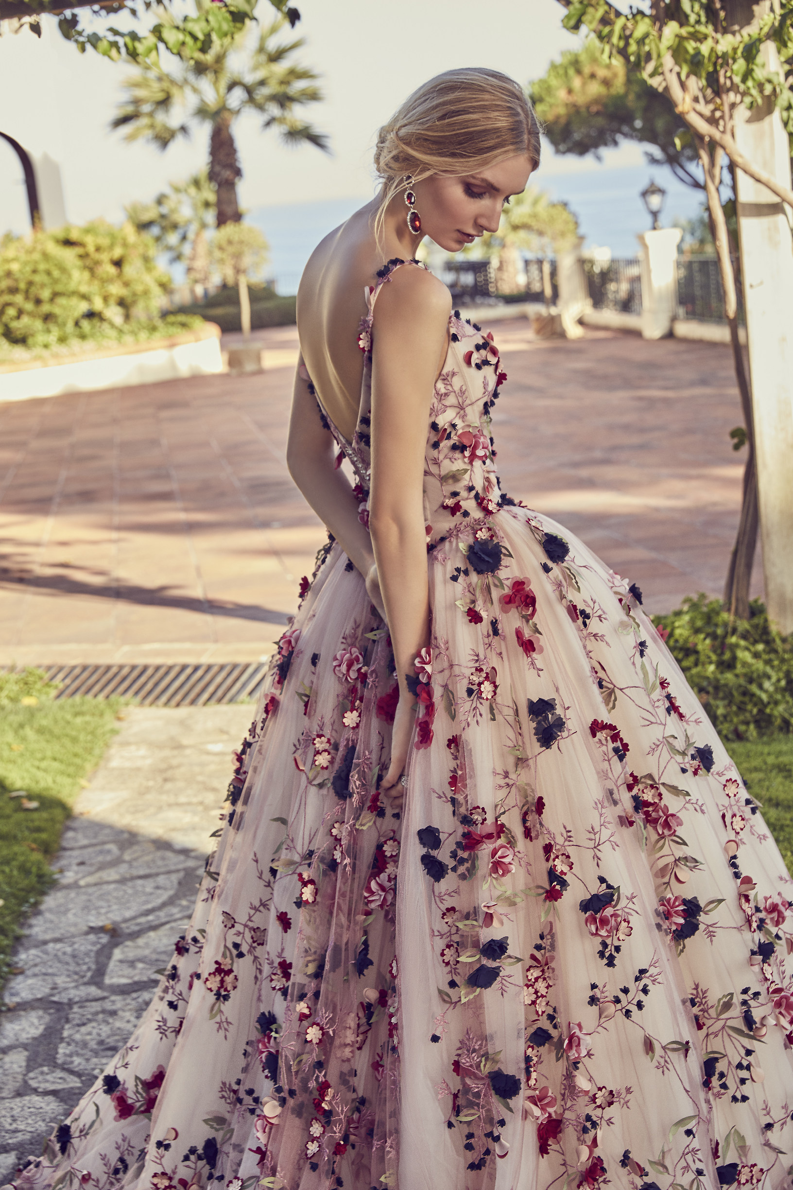 Model stood on a terrace near the sea wearing Ronald Joyce style 69426, a colourful boho wedding dress with navy, berry and green applique flowers and a ballgown skirt