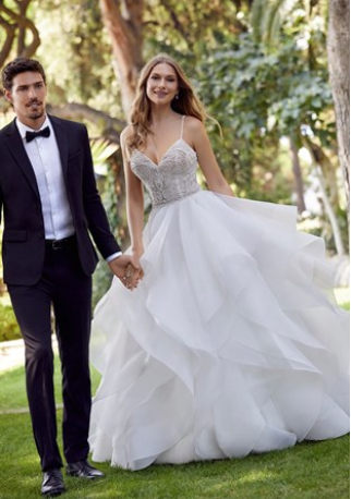 Model stood outside with a groom in Ronald Joyce style 69531, a princess ballgown wedding dress with a pattern beaded bodice, delicate straps and ruffled organza skirt