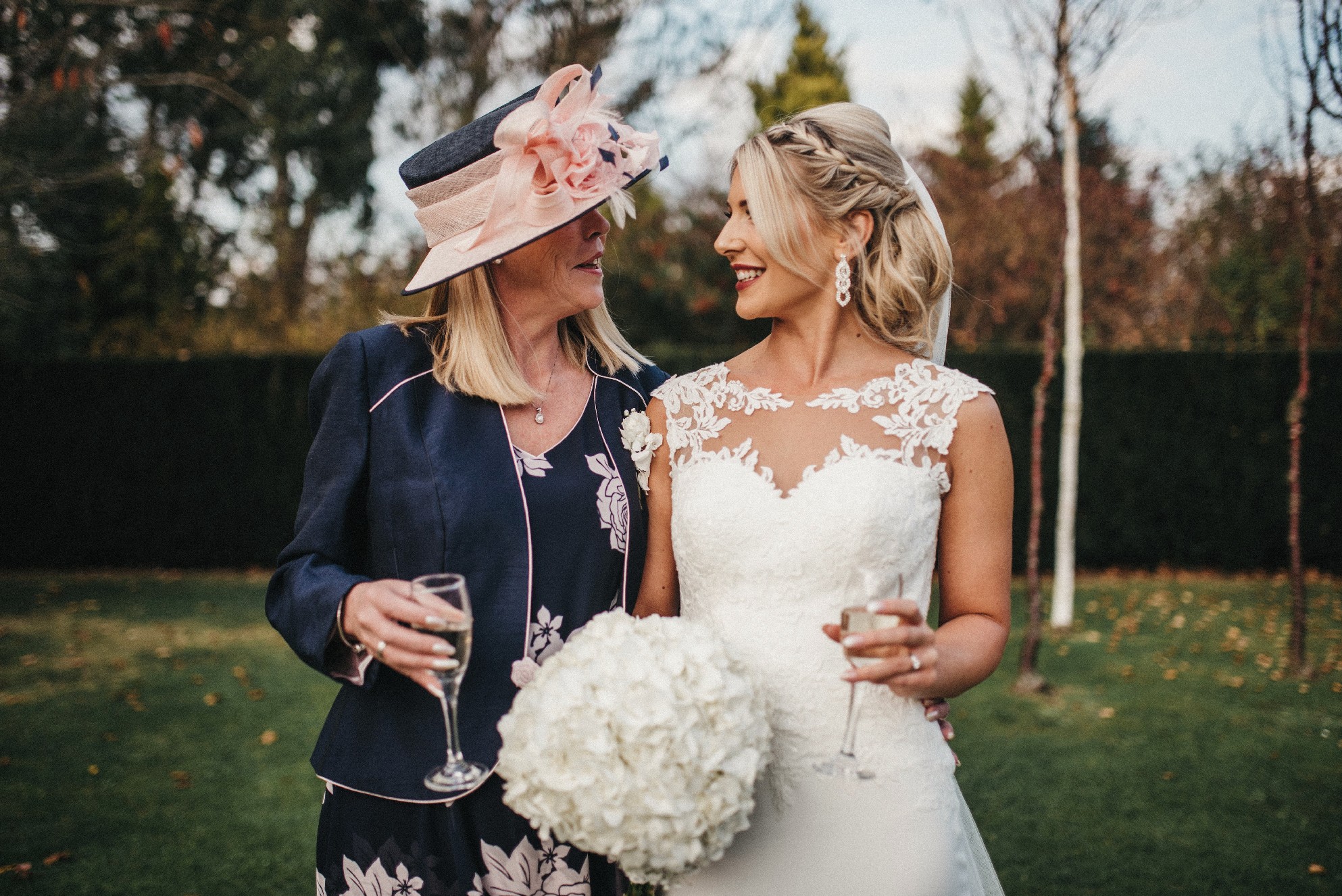 A photo of a bride stood next to her mum in a garden wearing an ivory sweetheart illusion wedding dress while holding a white bouquet and glass of fizz. 
