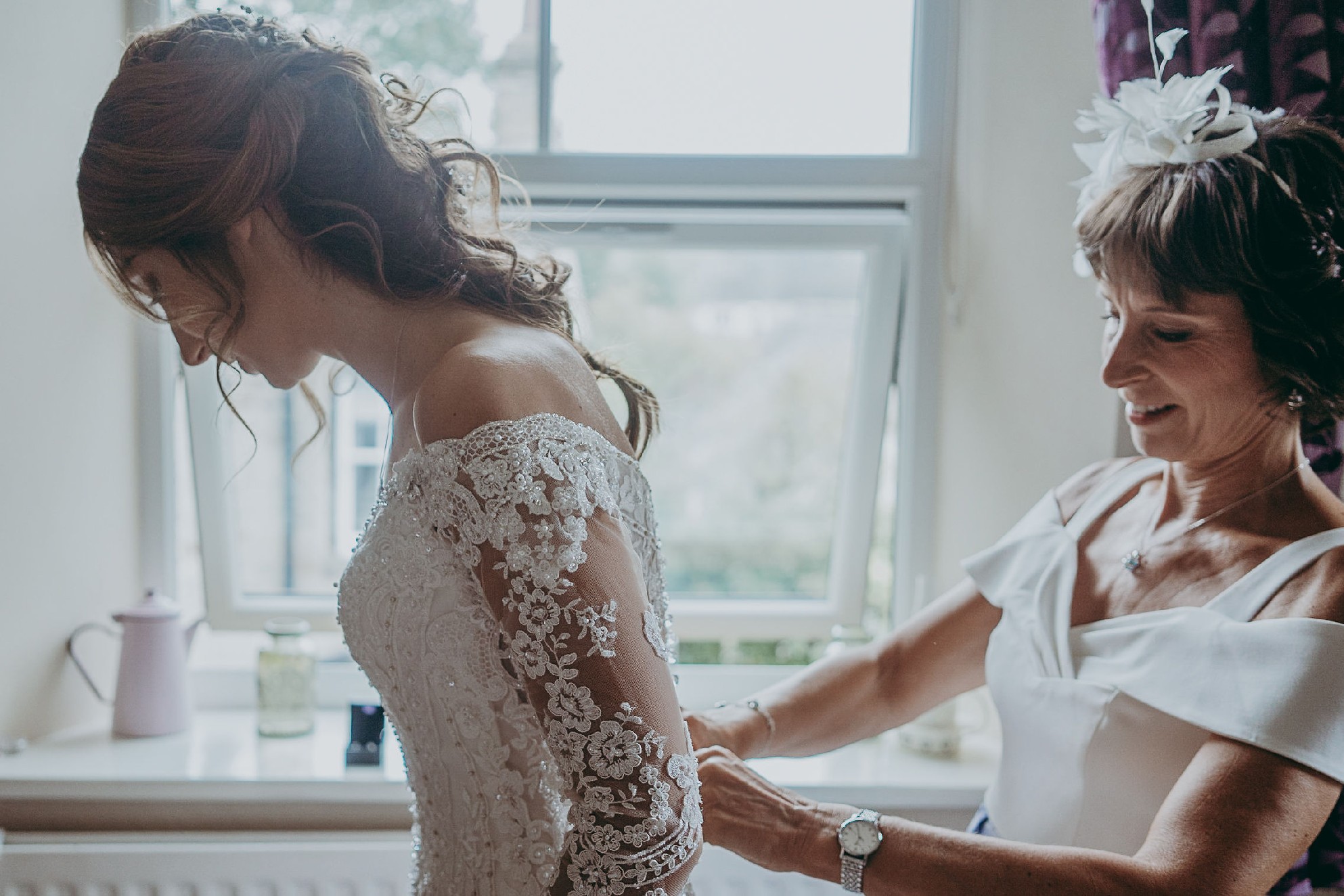 A close-up photo of a mum doing up the back of her daughter’s wedding dress on her wedding day. The bride wears an off the shoulder dress with long sheer and lace applique sleeves. 