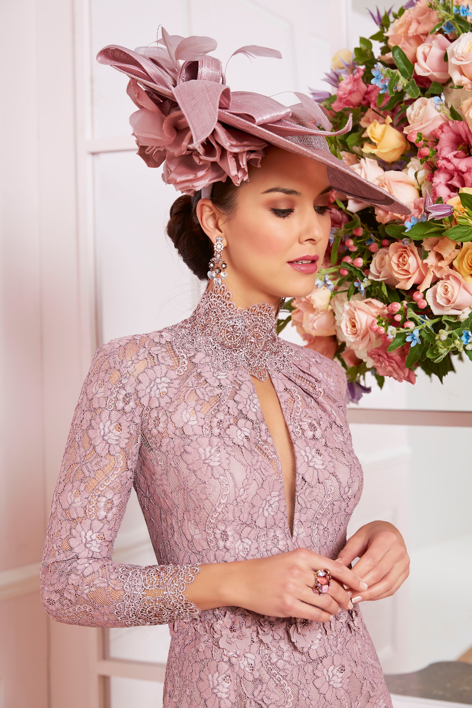 A brunette model wearing a dusty pink lace mother of the bride dress with a high neckline and keyhole detail. Model also wears a dusty pink statement hat. 
