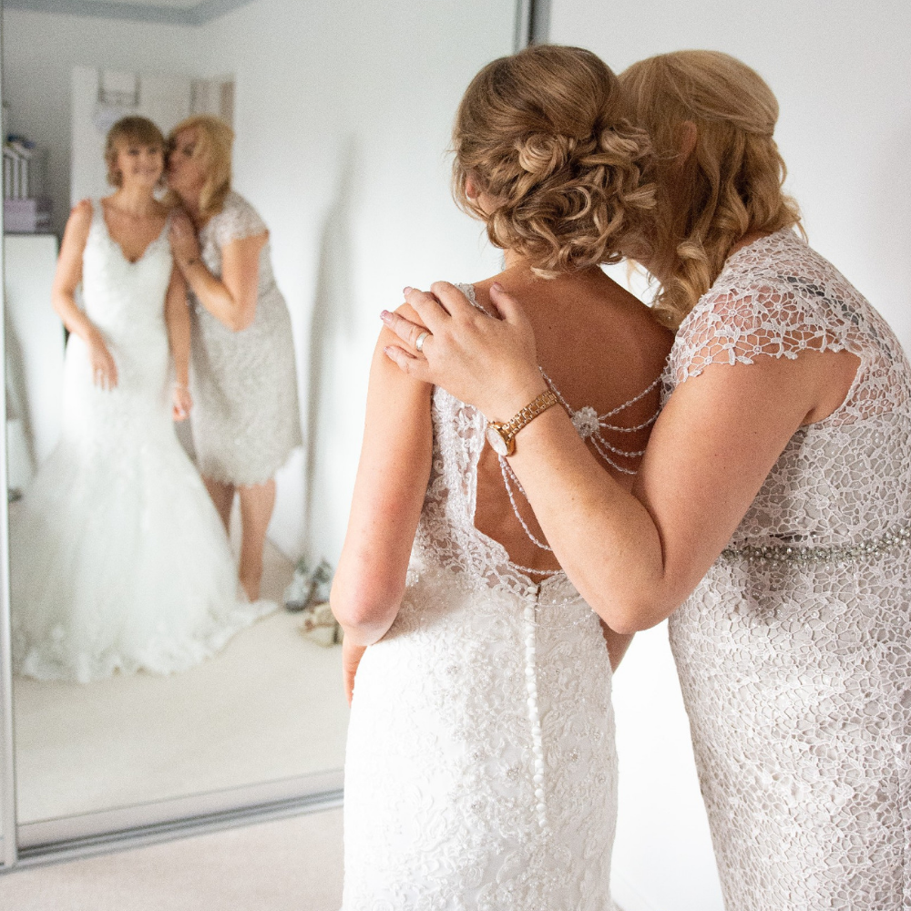 Rear profile photo of a bride stood next to her mum wearing a wedding dress while they both look at her reflection in a floor-length mirror