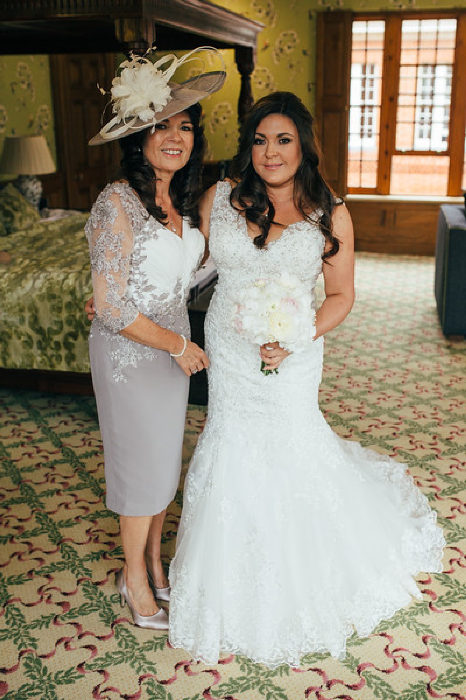 A photo of a bride stood with her mum near a mahogany four-poster bed in a hotel room. The bride wears a lace fishtail wedding dress with a v-neckline